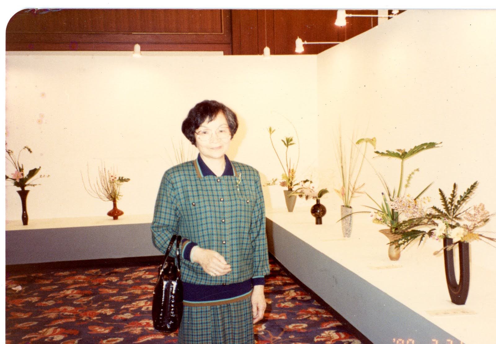 Image Description: Umi Hsu's Ahma wearing green and blue plaid jacket and skirt, gently smiling and standing in front of a row of ikebana arrangements on display in a gallery.