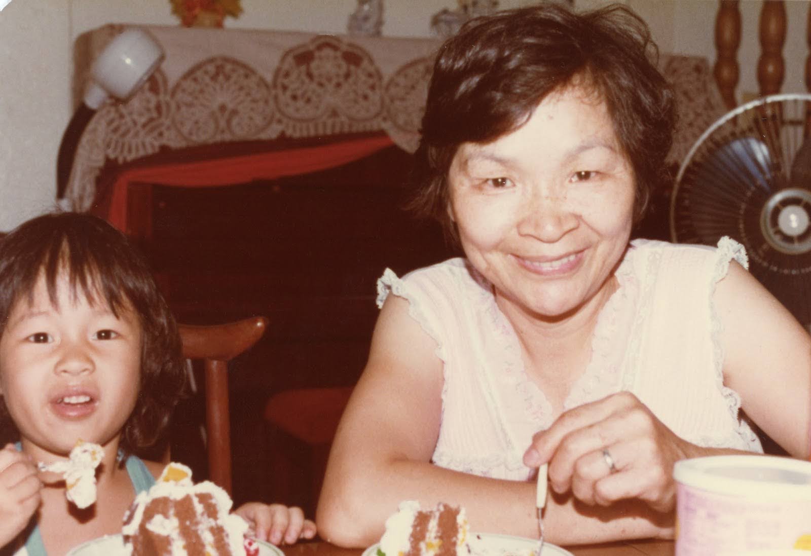 Image Description: Umi Hsu as a toddler eating cake next to Ahma wearing a white lacy tanktop. They are both smiling with forks of cake in hand. Behind them is a piano draped with cloth decoration. 